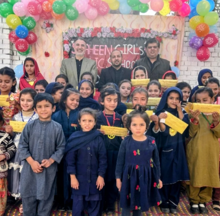 Children standing indoors, smiling and raising their hands, near colourful classroom posters.